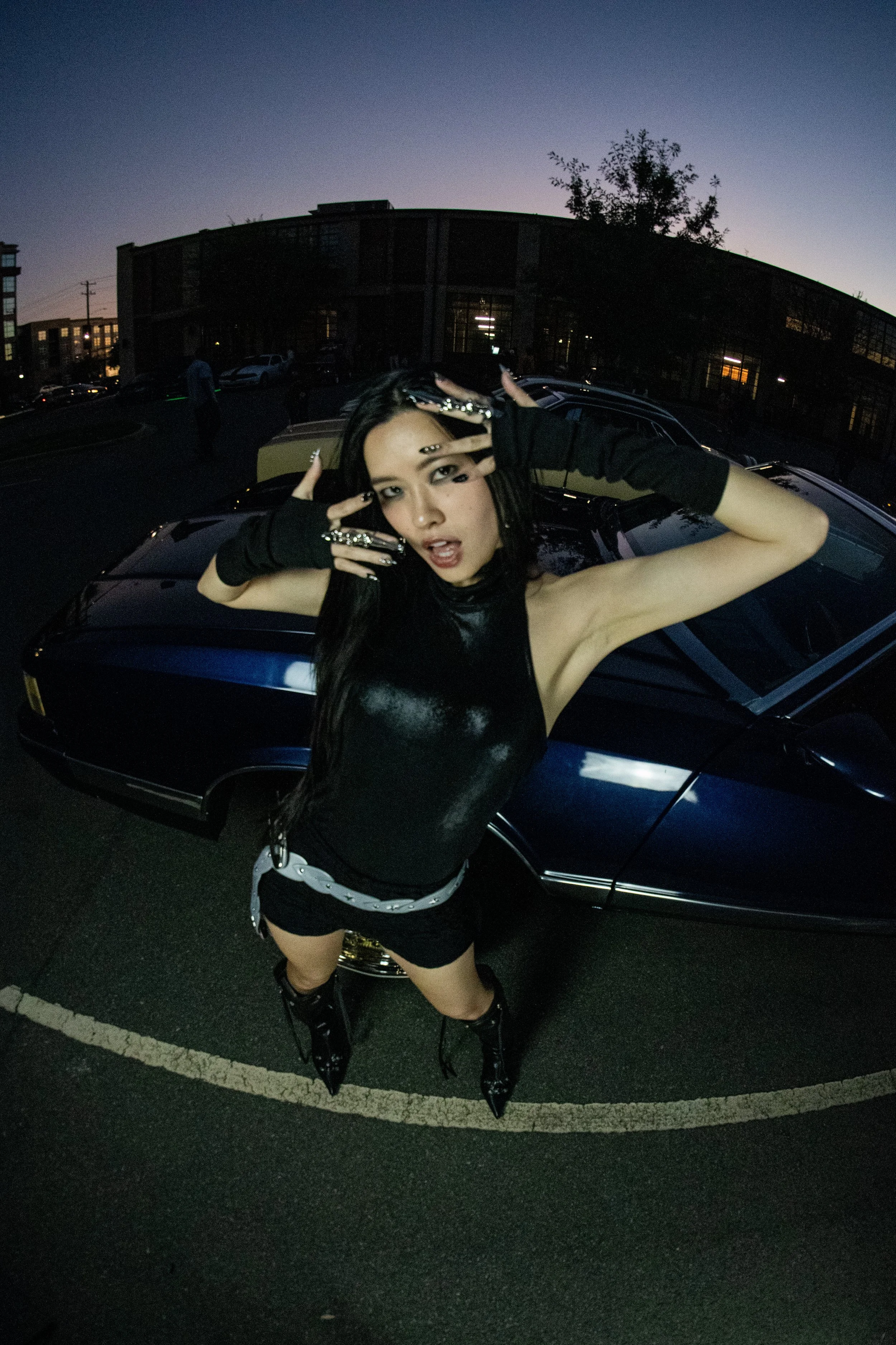 A woman with long dark hair, wearing black shiny sleeveless top, black shorts, and black high boots, poses with her hands near her face, standing in a parking lot at dusk, in front of a dark blue sports car.