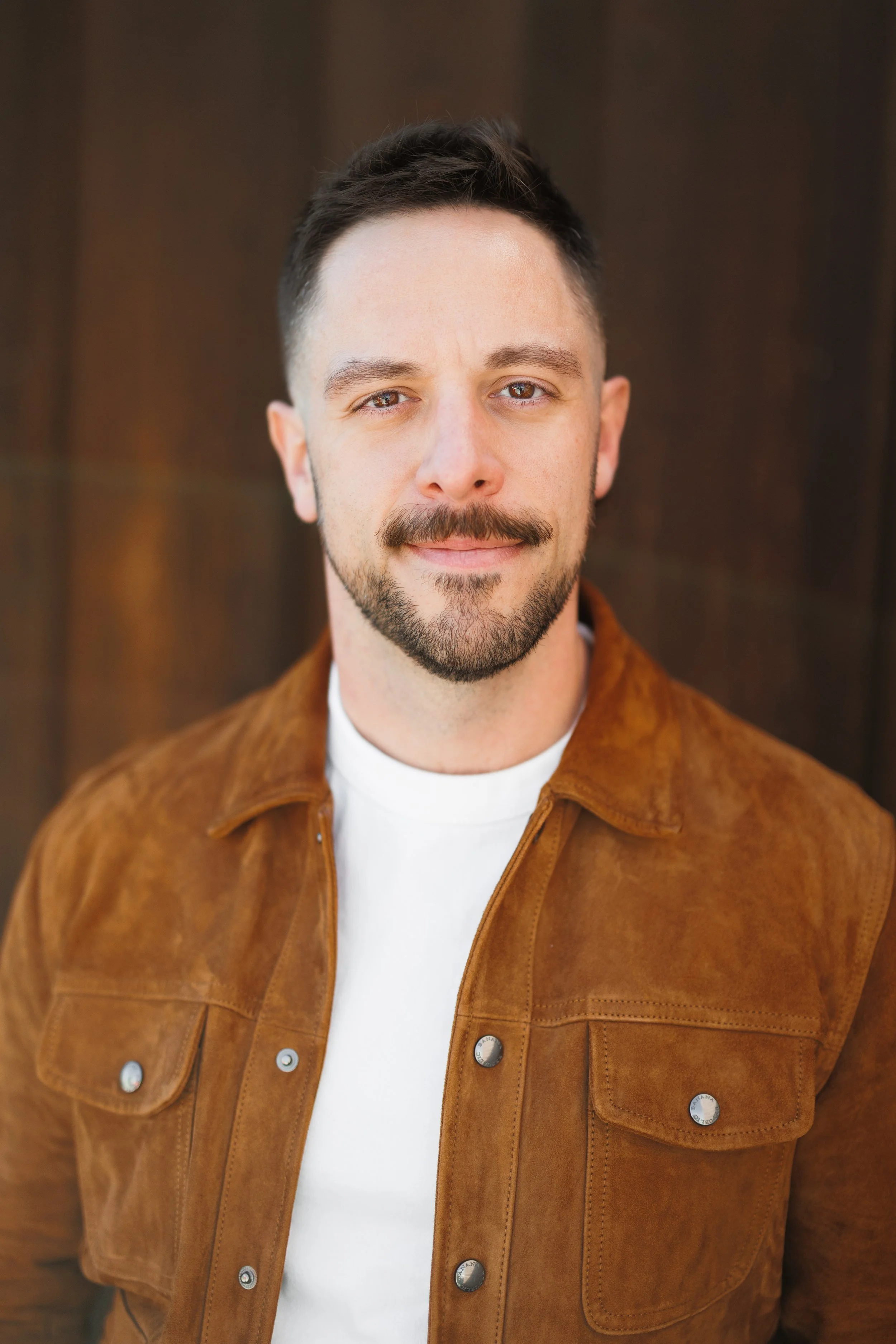 Close-up portrait of a man with short brown hair, light skin, brown eyes, a beard, and mustache, wearing a white t-shirt and brown suede jacket, standing against a blurred background.