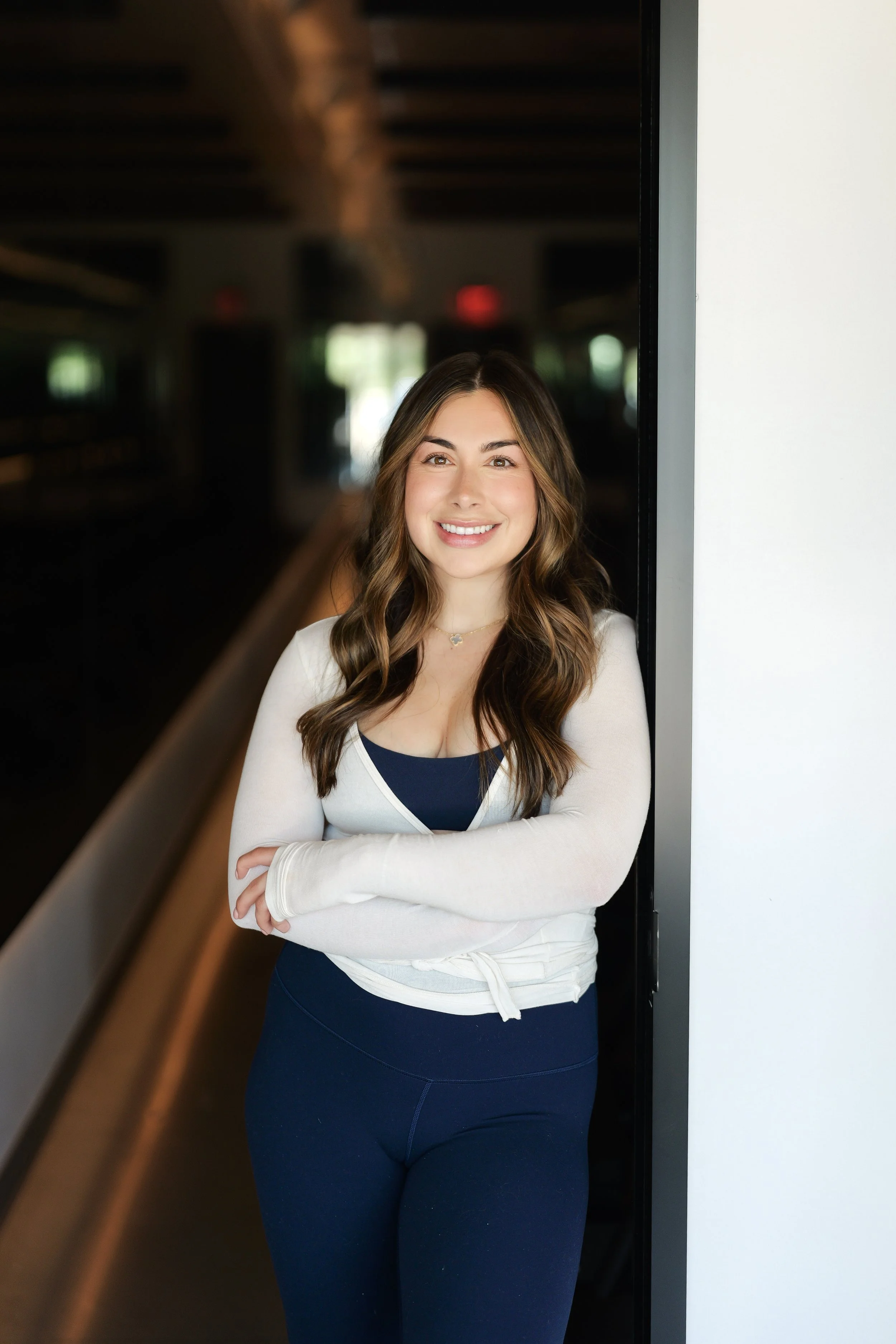 A young woman with long wavy hair, smiling, wearing a black long-sleeve top, standing against a dark background.