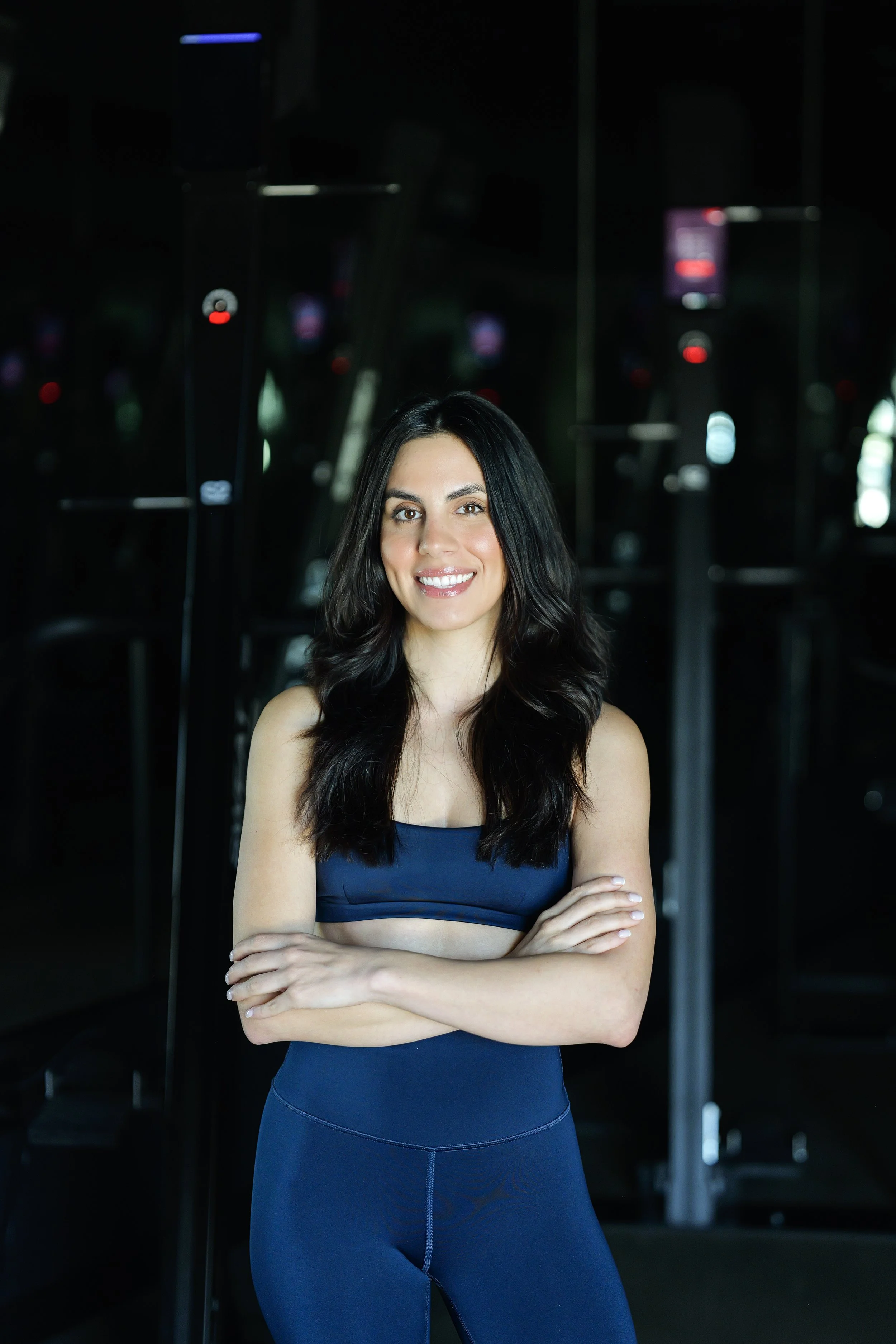 A woman with long dark hair, wearing a black sports bra and black leggings, stands with her arms crossed and smiles at the camera against a plain black background.