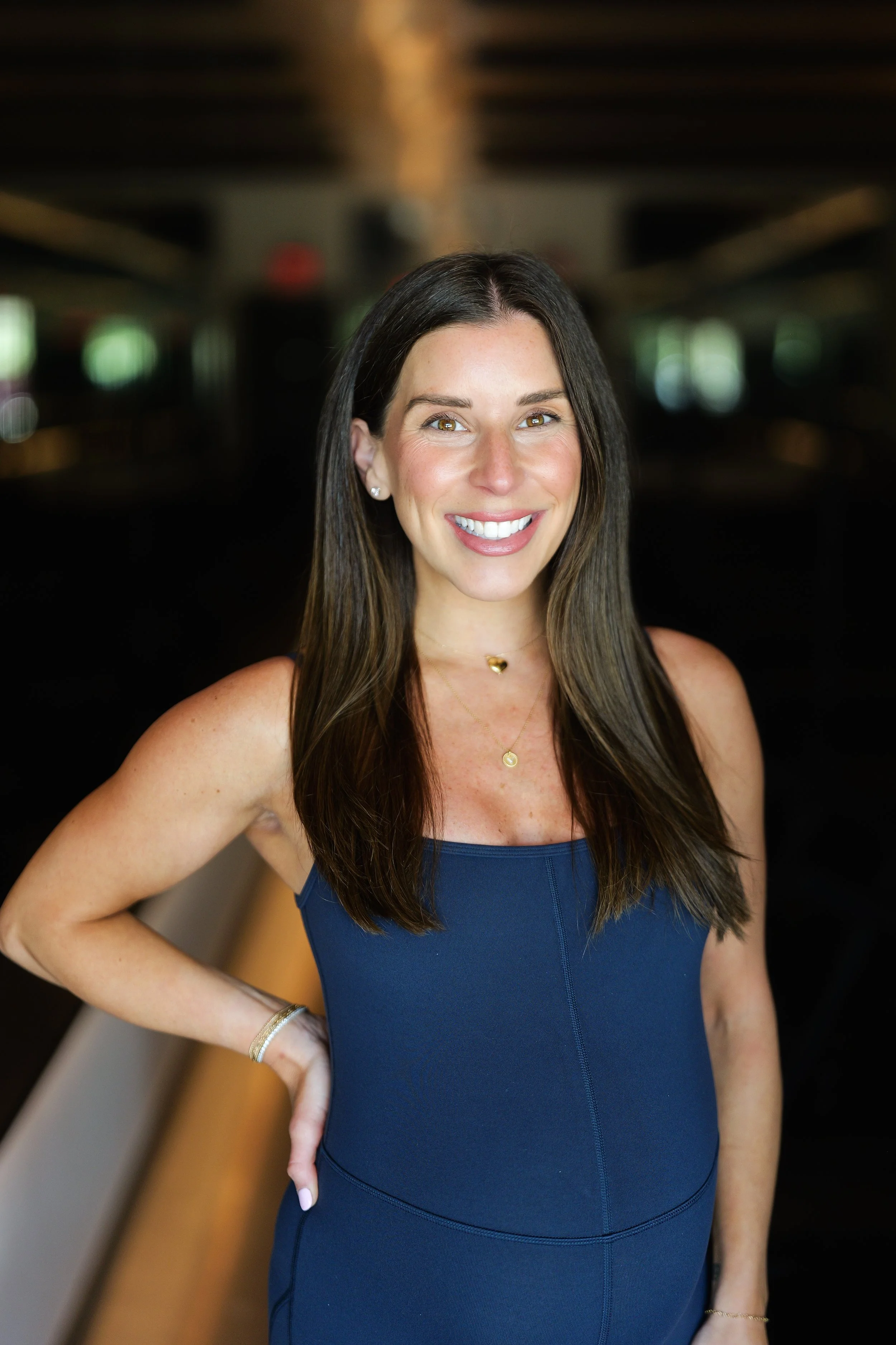 A smiling woman with long dark hair, wearing a black sports bra and black workout pants, standing with her hands on her hips against a dark background.