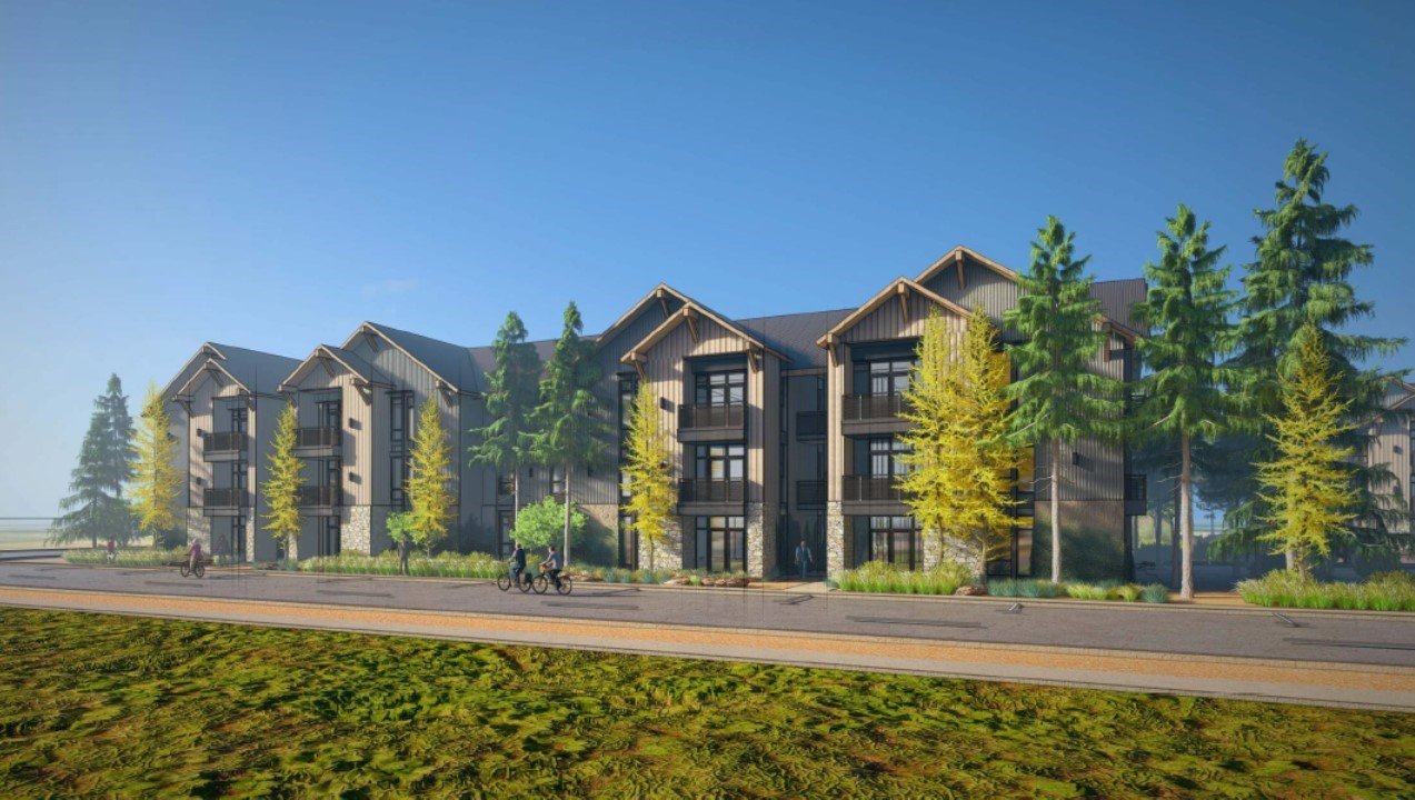 A modern multi-story apartment building with a dark exterior, surrounded by tall green trees and a clear blue sky. People are riding bikes on the street in front of the building.