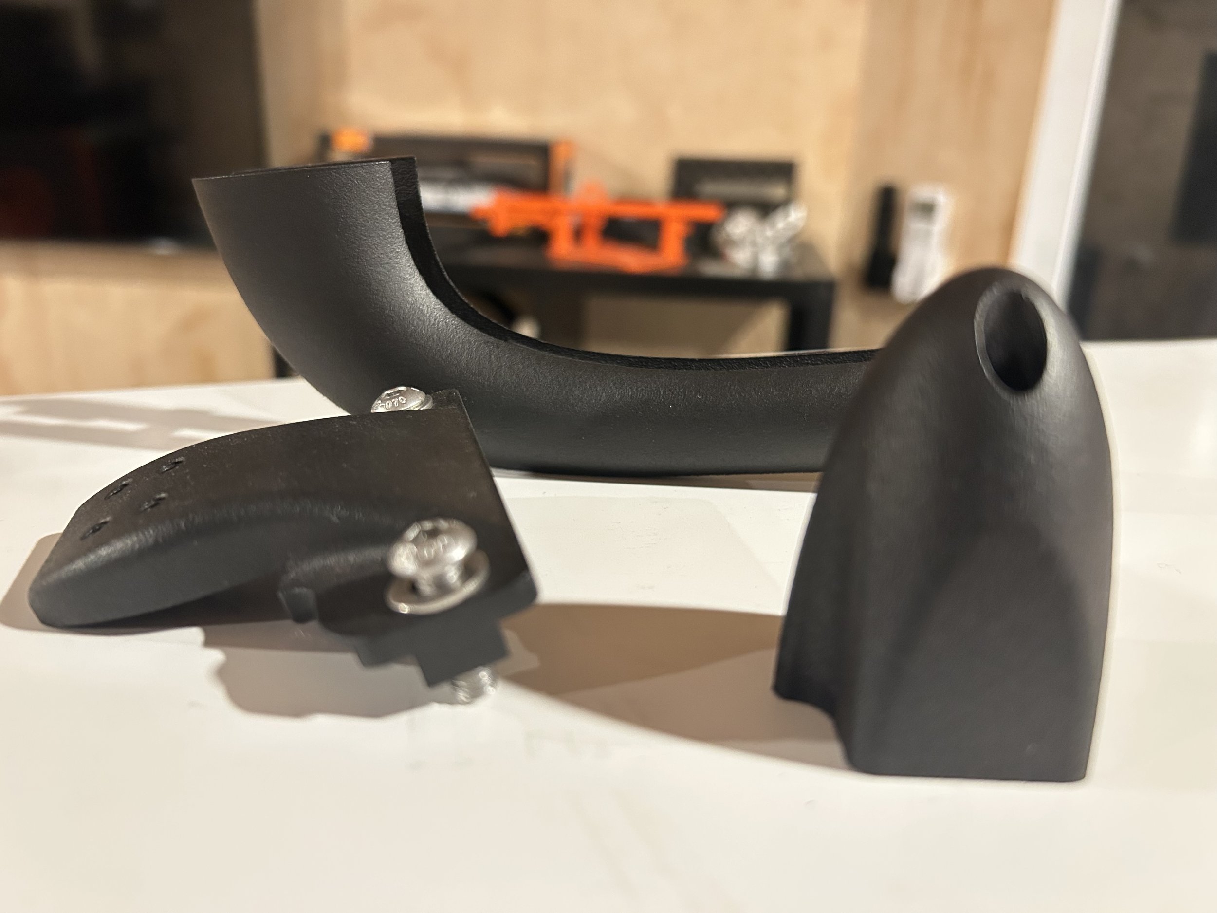 Close-up of a black plastic and metal bicycle handlebar stem. The stem has a hole at one end and bolts for attaching to a bicycle. The background includes a blurred room with furniture and tools.