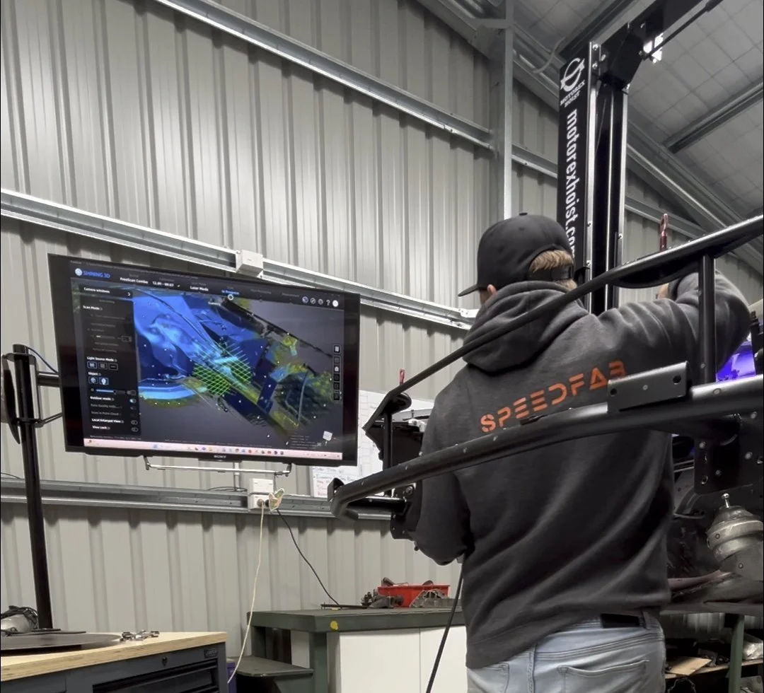 A person wearing a black cap and gray hoodie with orange letters working on a racing car inside a workshop. The workshop has metal walls and a monitor displaying a 3D model of the car on a stand.