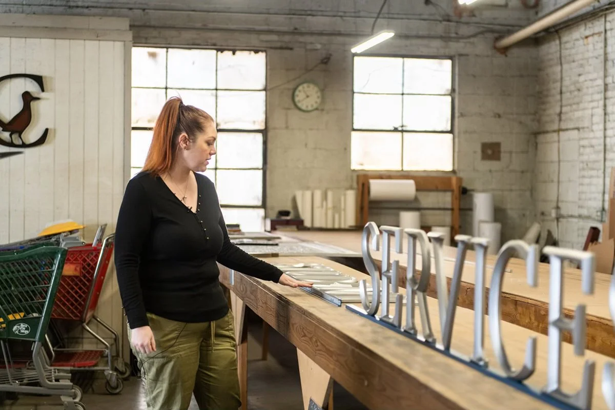 A woman inspects a metal sign that spells 'SERVICES' on a workbench inside a foundry workshop, with industrial tools and materials in the background. The space features large windows and high ceilings, giving it an authentic, industrial feel.