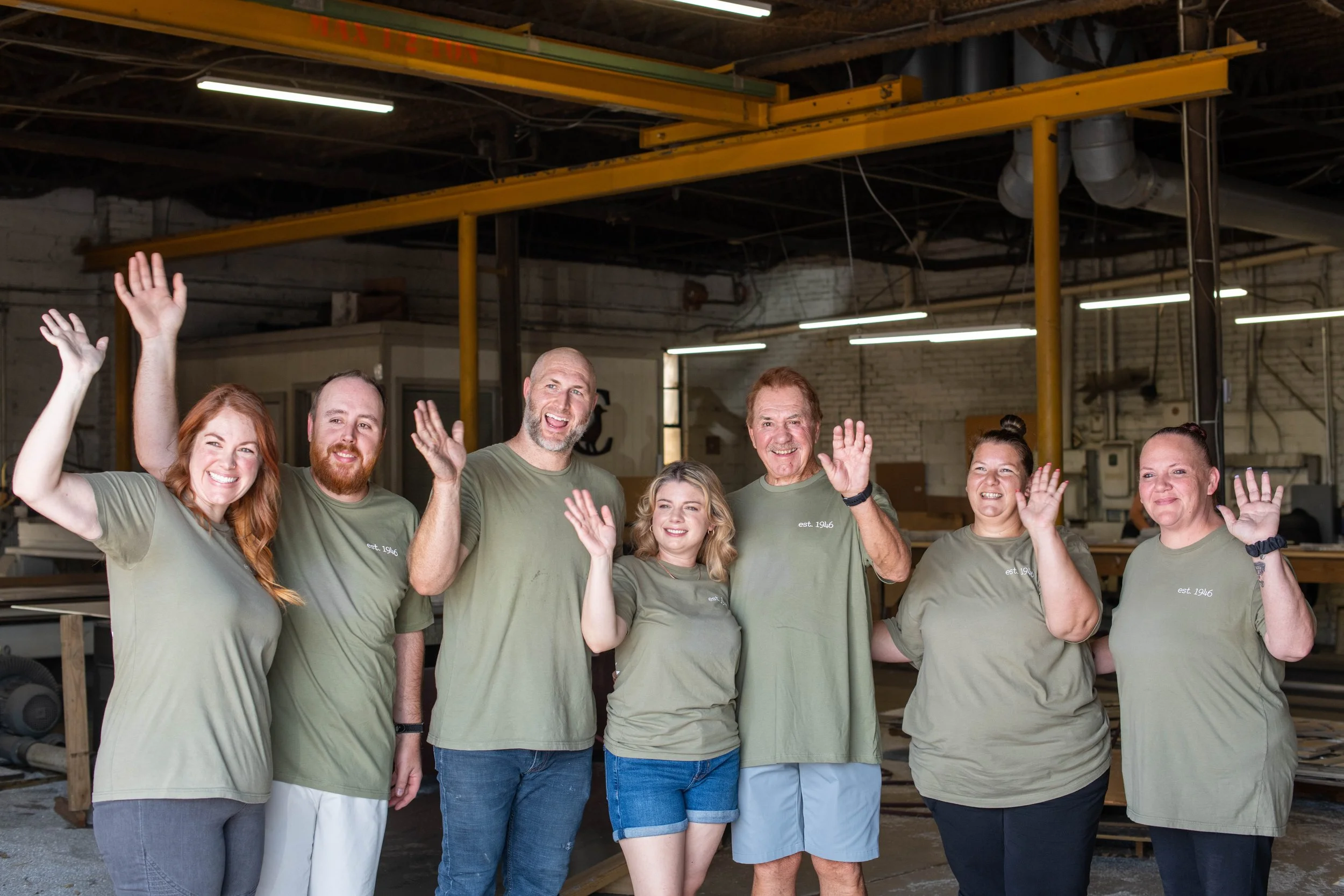 Smiling A.R.K. Ramos team waving together outside the foundry, celebrating their legacy as a family owned American-made signage company.