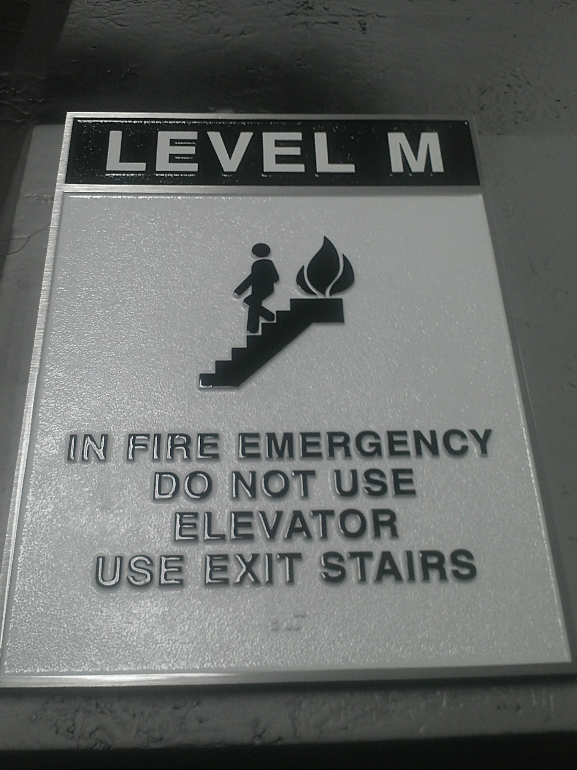 ADA signage with tactile lettering and fire safety instructions reading “In Fire Emergency Do Not Use Elevator Use Exit Stairs,” showcasing compliant wayfinding by A.R.K. Ramos.