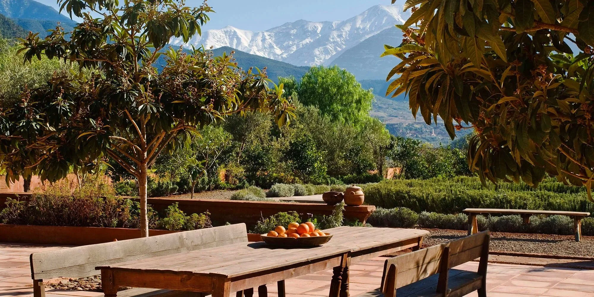 Outdoor patio with wooden table and chairs, bowl of peaches, lush green trees, and mountains in the background.