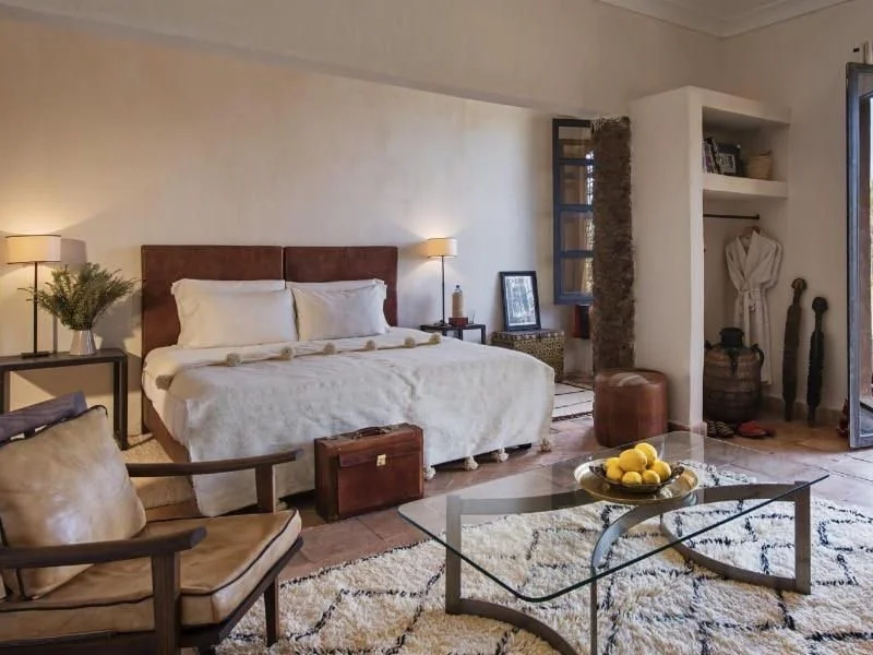 A cozy bedroom featuring a king-sized bed with a brown headboard, white bedding, and pillows, flanked by two lamps. There is a leather armchair, a glass coffee table with a bowl of lemons, a small side table with a plant, and a built-in open closet with robes and decorative items. A window and a door lead to an outdoor space, and the room has a rustic, warm decor with wooden accents and textured walls.
