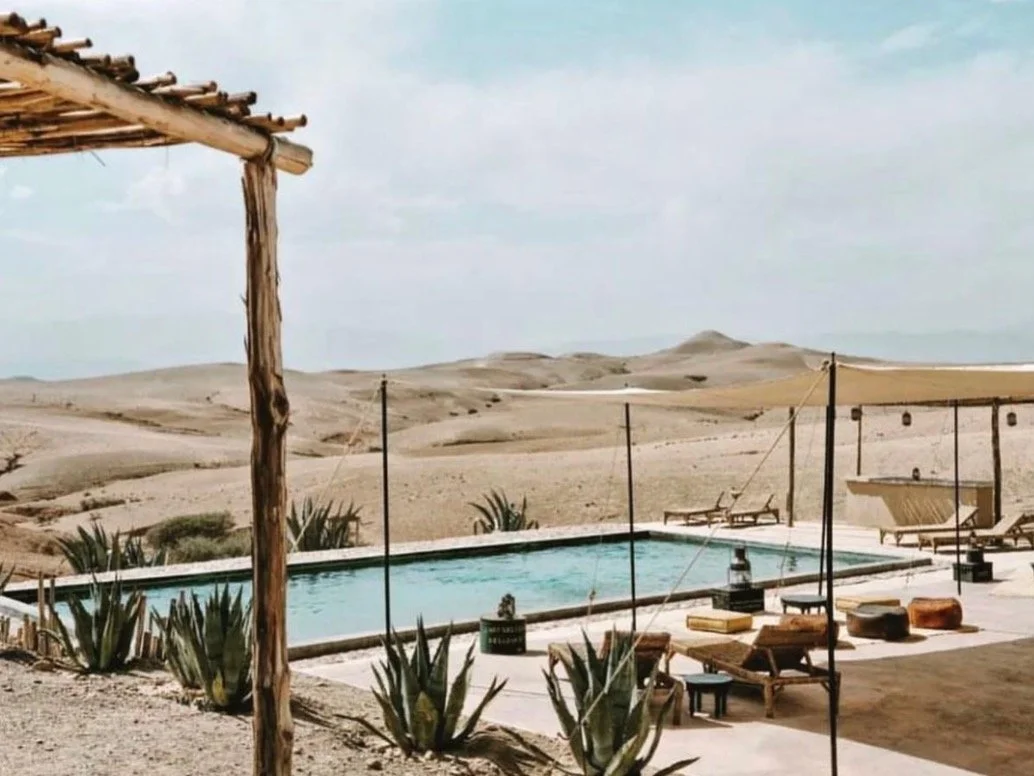 Outdoor pool surrounded by desert landscape with sand dunes, sun loungers, and sun umbrellas.