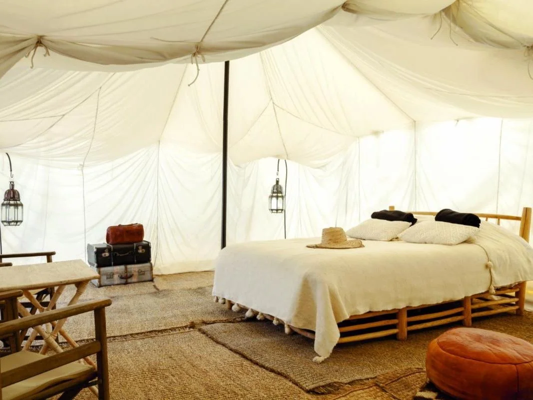 A cozy camping tent with a bed, pillows, a hat on the bed, and wooden furniture including chairs and a table, with lanterns hanging inside.
