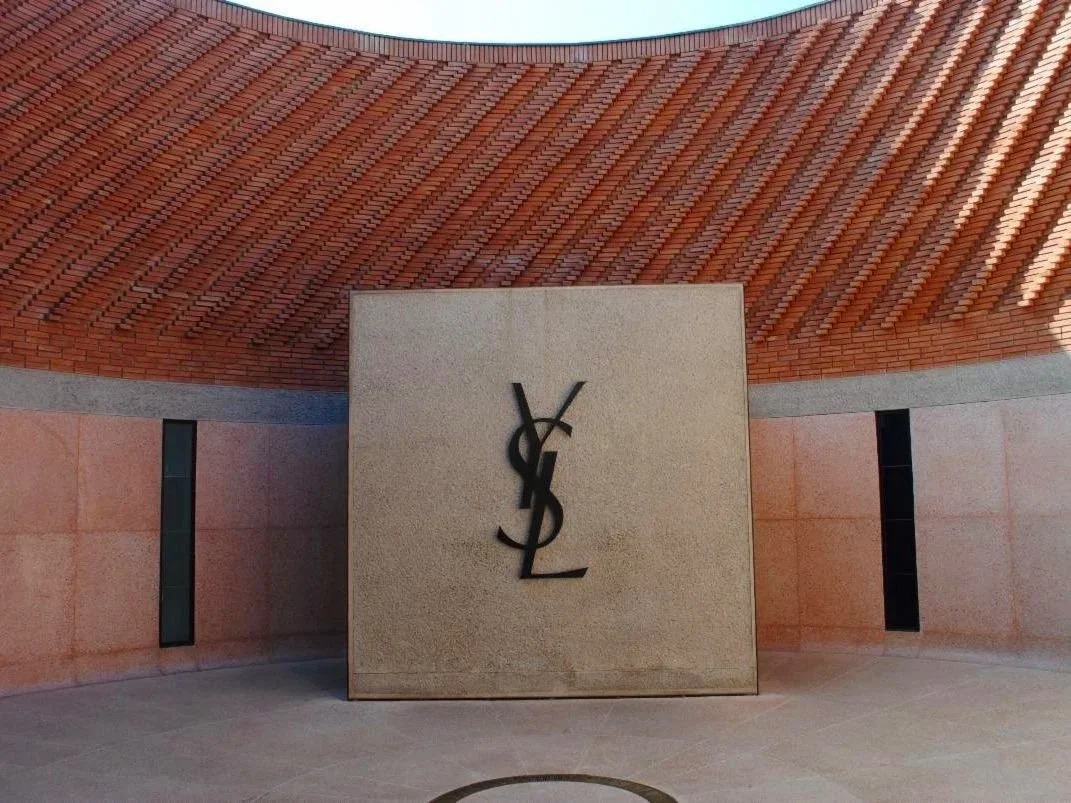 A modern building with a curved red brick roof and pinkish walls with narrow vertical windows. In front, there is a beige stone wall with a black Yves Saint Laurent logo.