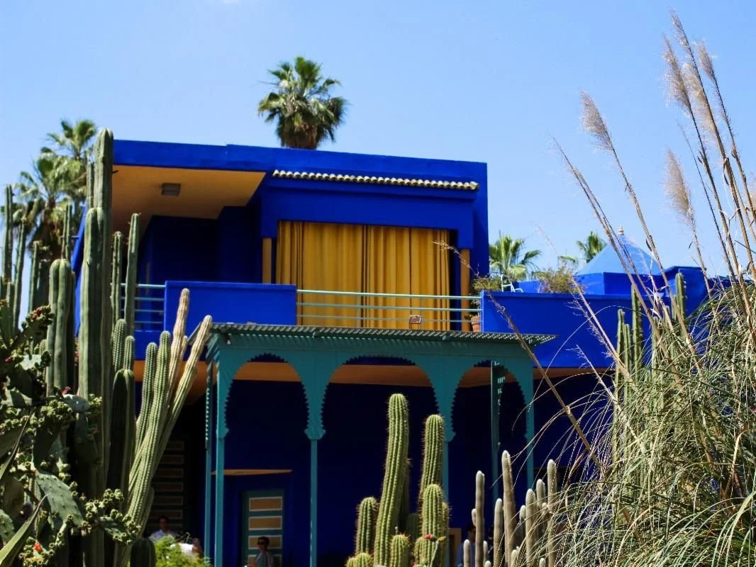 Bright blue modern house with yellow curtains, surrounded by tall cacti and desert plants, under a clear blue sky, Jardin Majorelle.