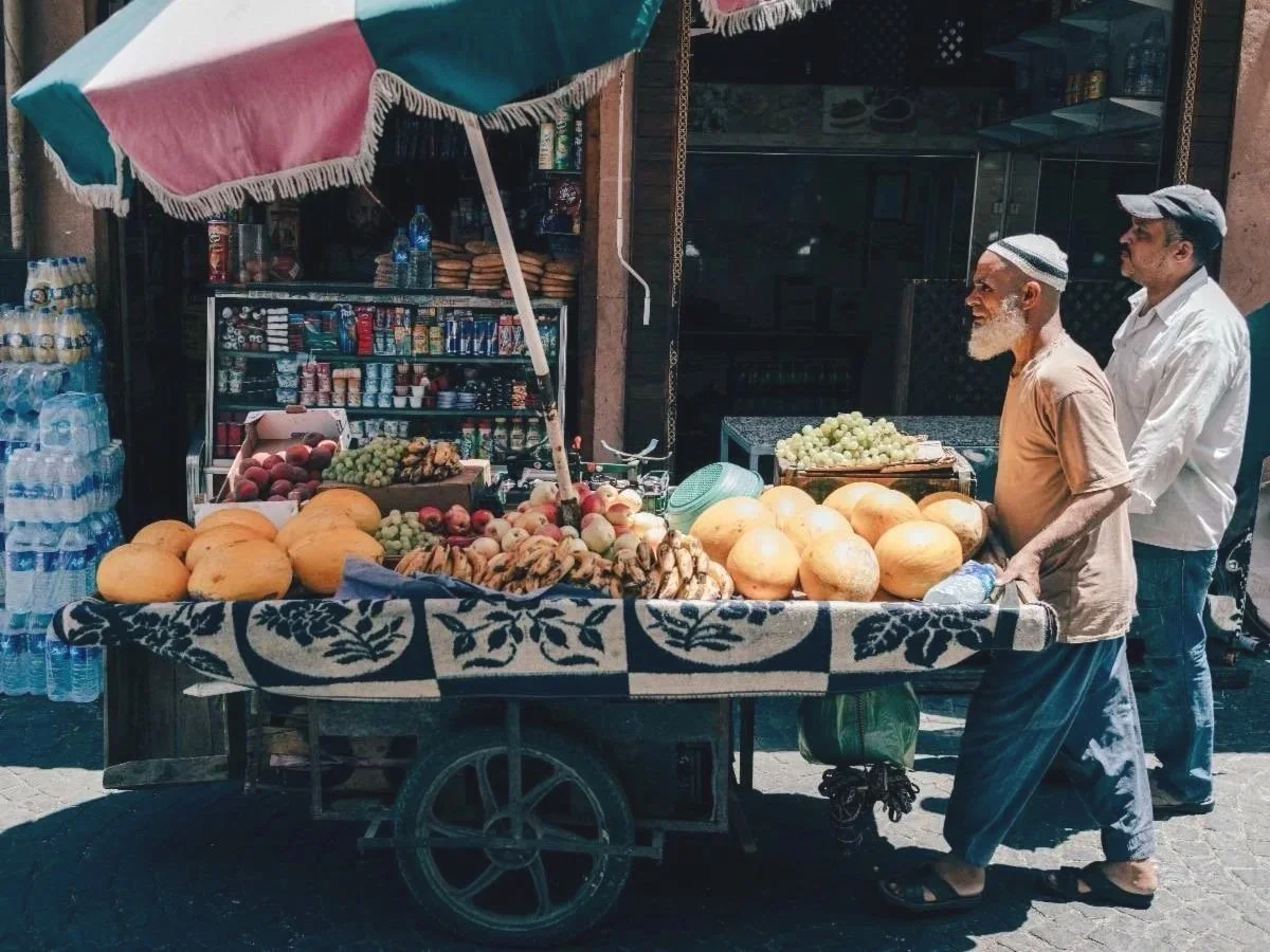 A street vendor's cart filled with melons, grapes, peaches, and bananas, with a shaded umbrella and a small store selling snacks and bottled water behind.