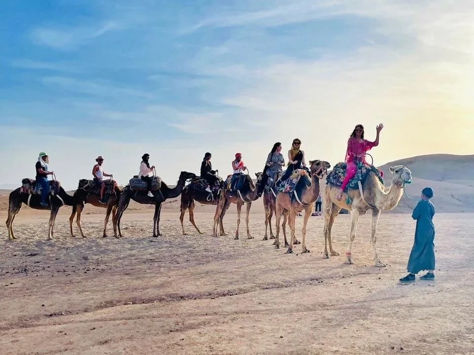 People riding camels in a desert with a guide walking beside them, under a blue sky with some clouds.