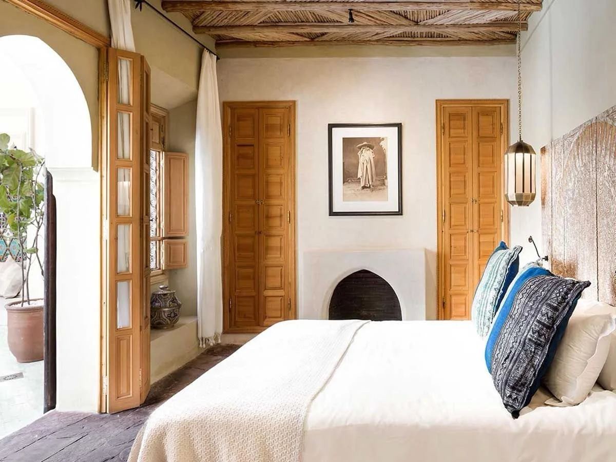 A cozy bedroom with a white bed, colorful pillows, wooden accents, a framed picture on the wall, and a hanging lamp. An open door reveals a potted plant outside.