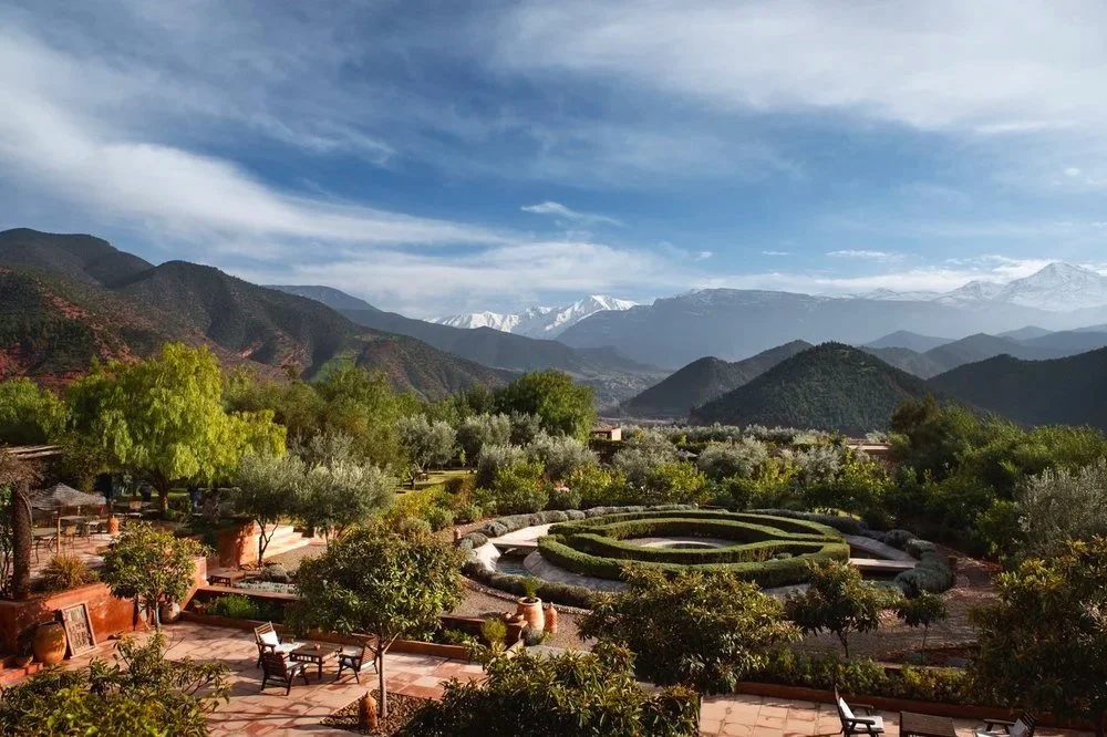 A scenic mountain landscape with lush green trees, a circular garden, and a patio with benches in the foreground.