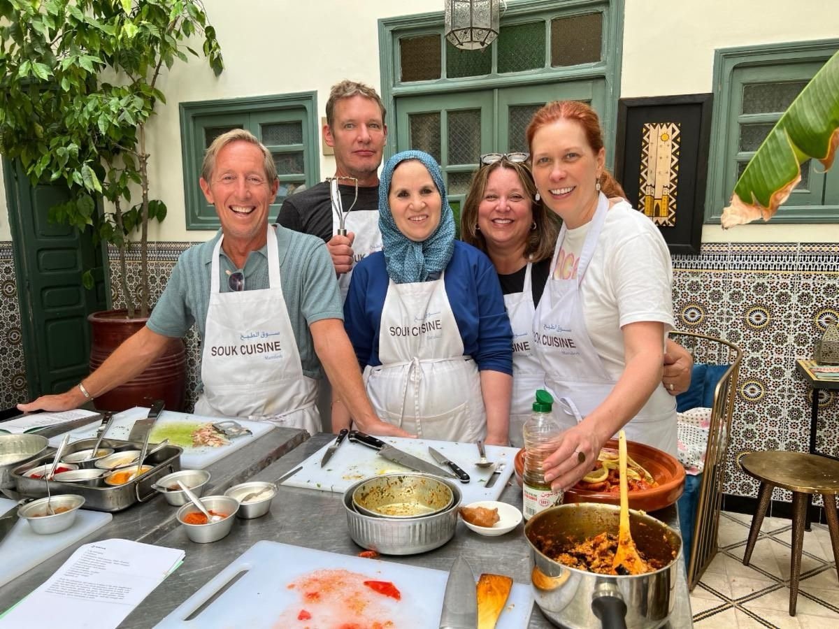 Group of five people wearing aprons in a kitchen, smiling for a photo, with cooking ingredients and utensils on the table in front of them.