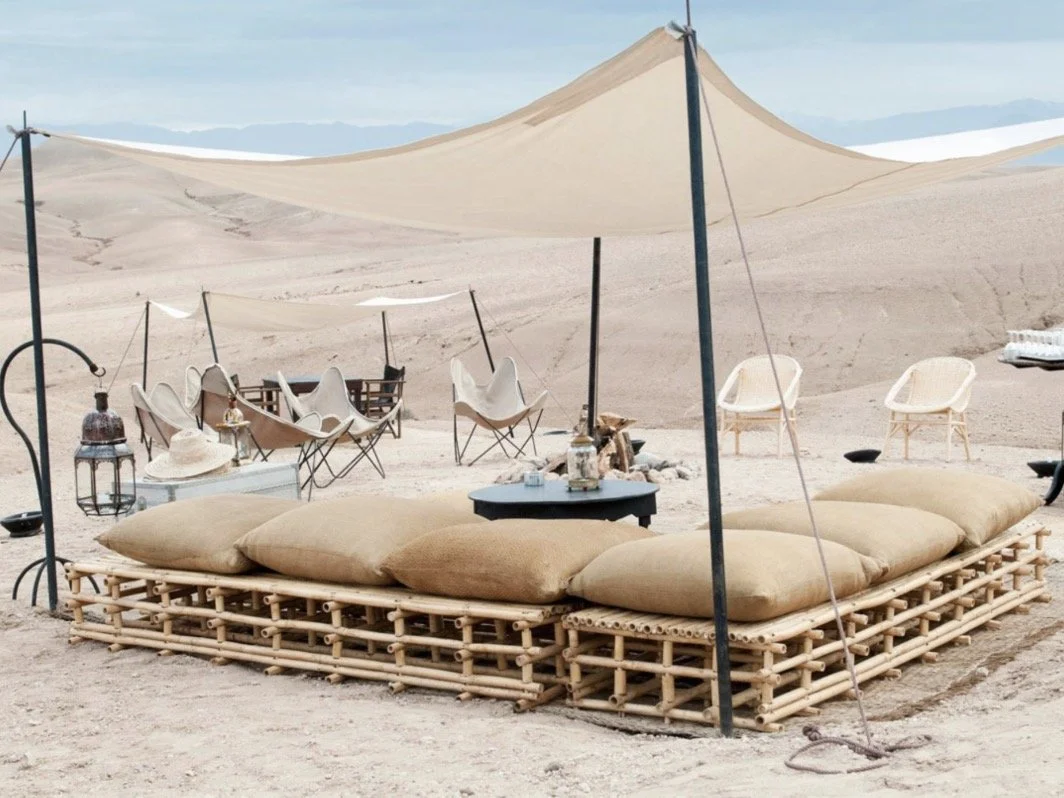 A desert setting with a large bamboo daybed with beige cushions under a beige canopy, surrounded by chairs, lanterns, and other decorative items.