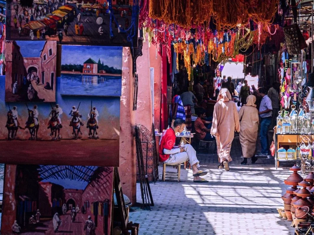 Market scene with people walking and shopping, hanging decorations, paintings on display, and various items for sale, including pottery and jewelry.