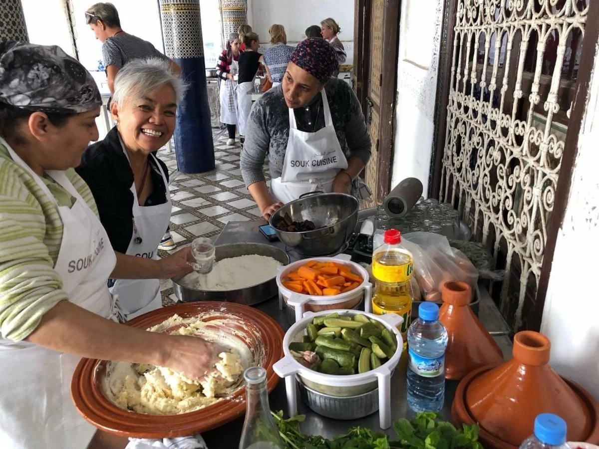 Group of women cooking together in a kitchen with traditional Moroccan tagines, vegetables, and cooking supplies.
