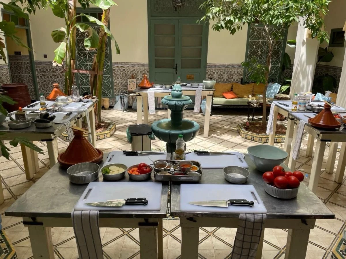Indoor Moroccan-style dining area with tables set with plates, knives, and bowls, and a central fountain surrounded by plants and decorative tiles.