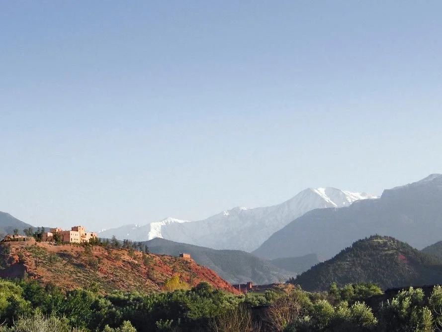 Mountain landscape with snowy peaks in the background, desert hills with buildings, and green vegetation in the foreground.