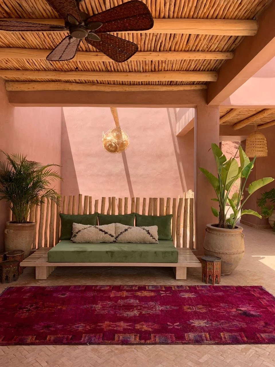 A cozy indoor space with a wooden ceiling and large potted plants flanking a green cushioned seating area. A red patterned rug covers the floor, and a woven pendant light hangs from the ceiling.