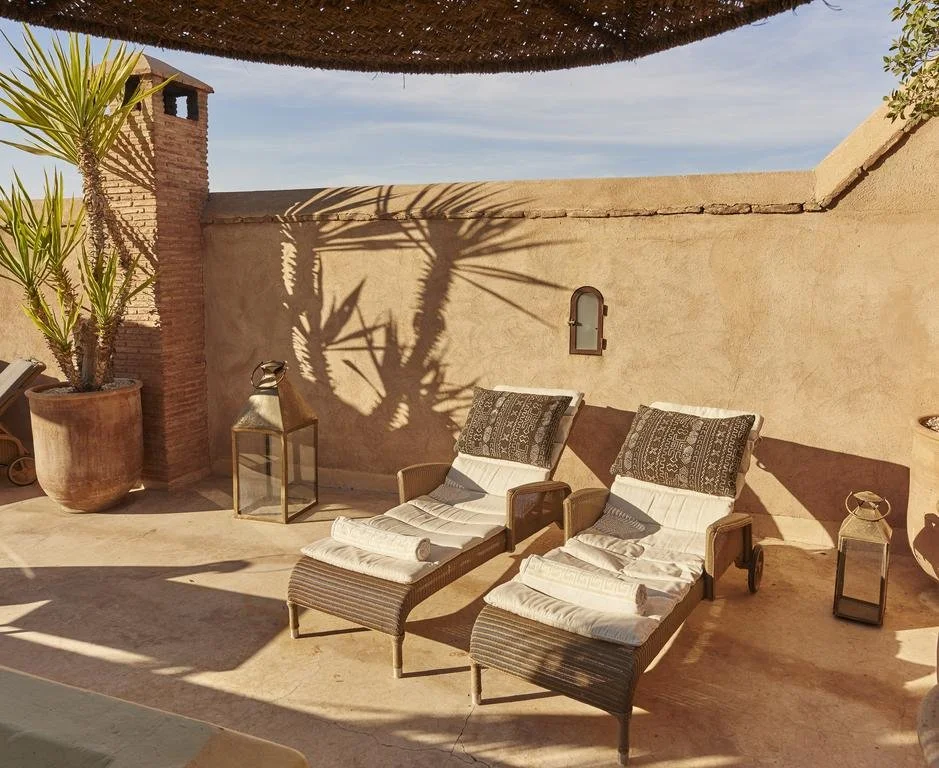 Outdoor patio with two lounge chairs with cushioned pillows, potted plants, lanterns, and a shaded area, casting shadows on a beige wall.