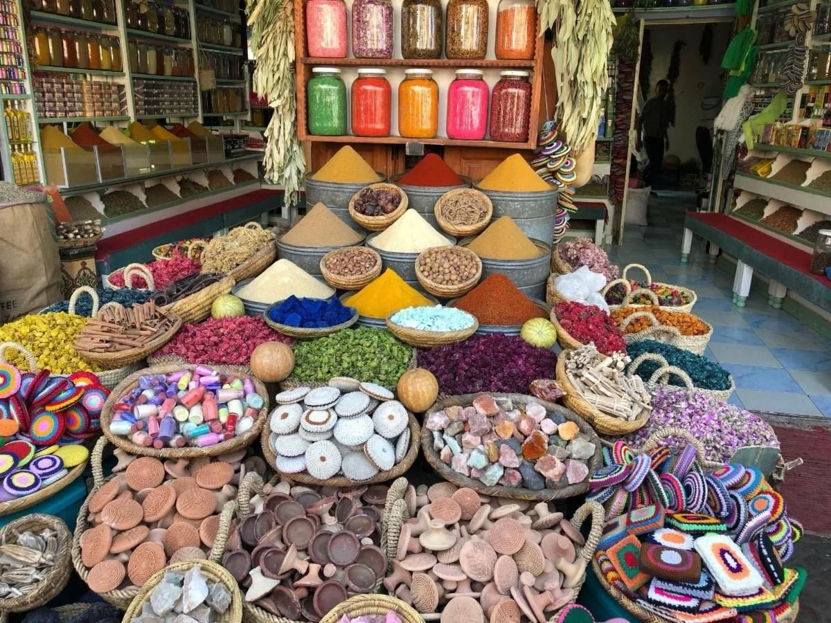 A colorful marketplace display of various spices, herbs, and herbs in baskets, with jars of powders above and shelves of products in the background.