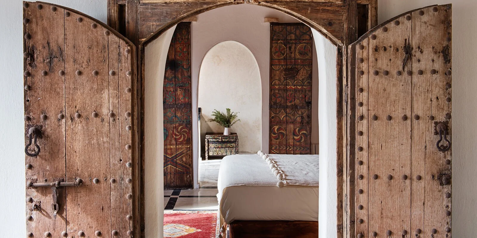View through rustic wooden doors into a bedroom with a white bed, colorful painted doors, a black and white patterned rug, and a small table with a plant in a white vase.