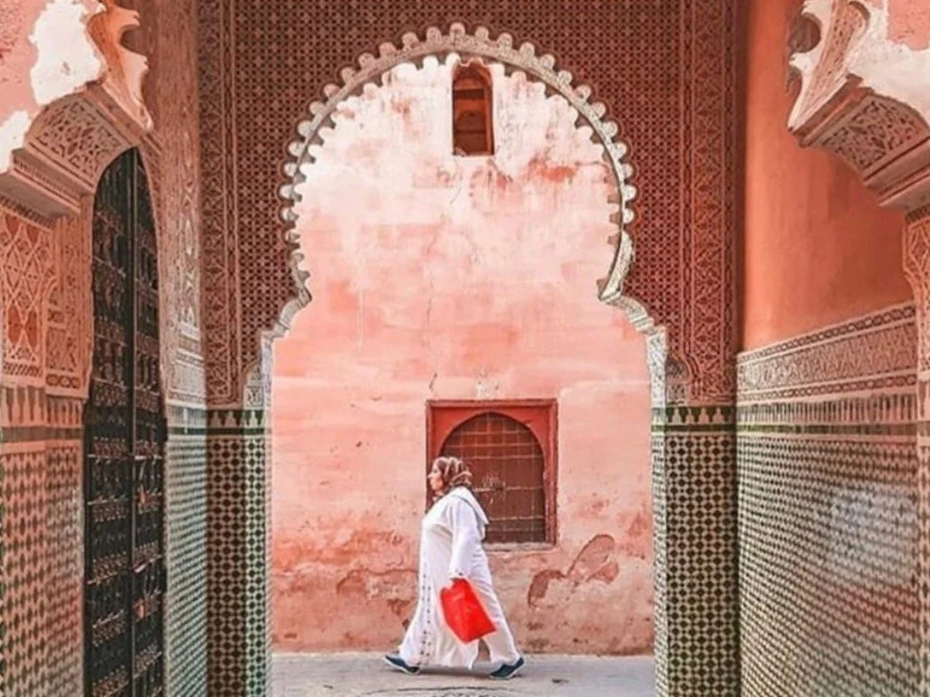A person walking inside a corridor of a historic building with intricate architectural details, arched doorways, and colorful tile work.