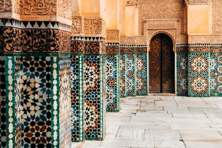 Decorative mosaic Moroccan tiles on walls and columns inside a building with a wooden door and stone floor.