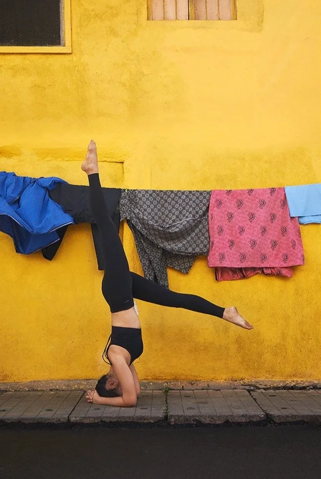 Femme faisant un équilibre en position de tête sur la tête, dans un style de yoga, devant un mur jaune avec des vêtements suspendus.