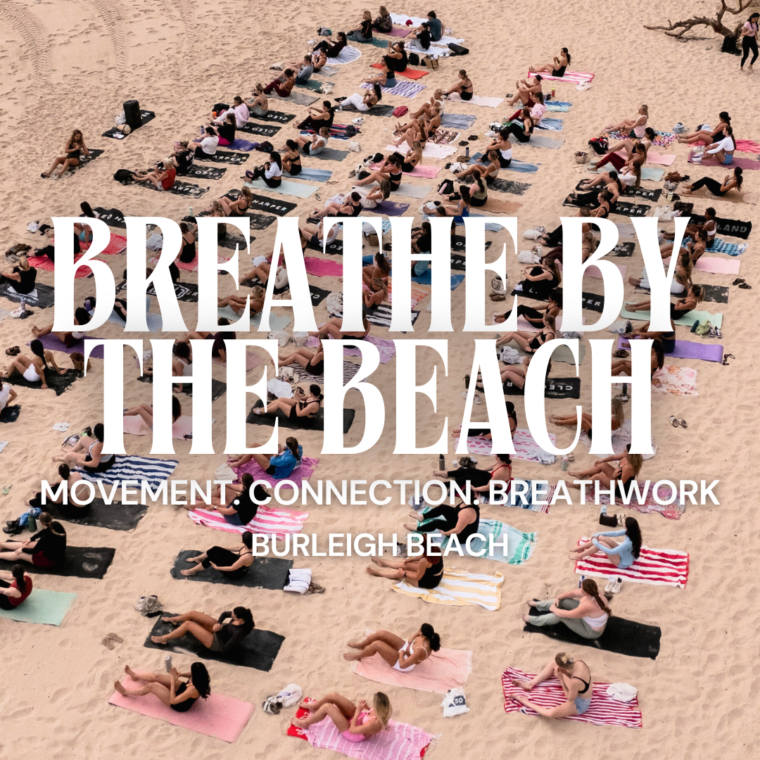 Aerial view of people practicing yoga on a sandy beach, with a group sitting in rows on yoga mats near the water, and a larger group standing in the distance.