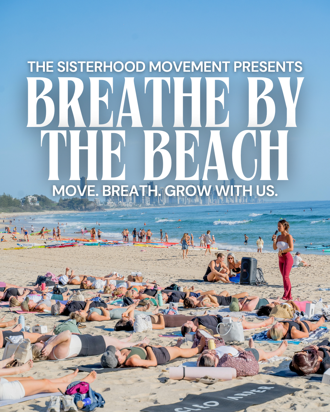 Aerial view of people practicing yoga on a sandy beach, with a group sitting in rows on yoga mats near the water, and a larger group standing in the distance.