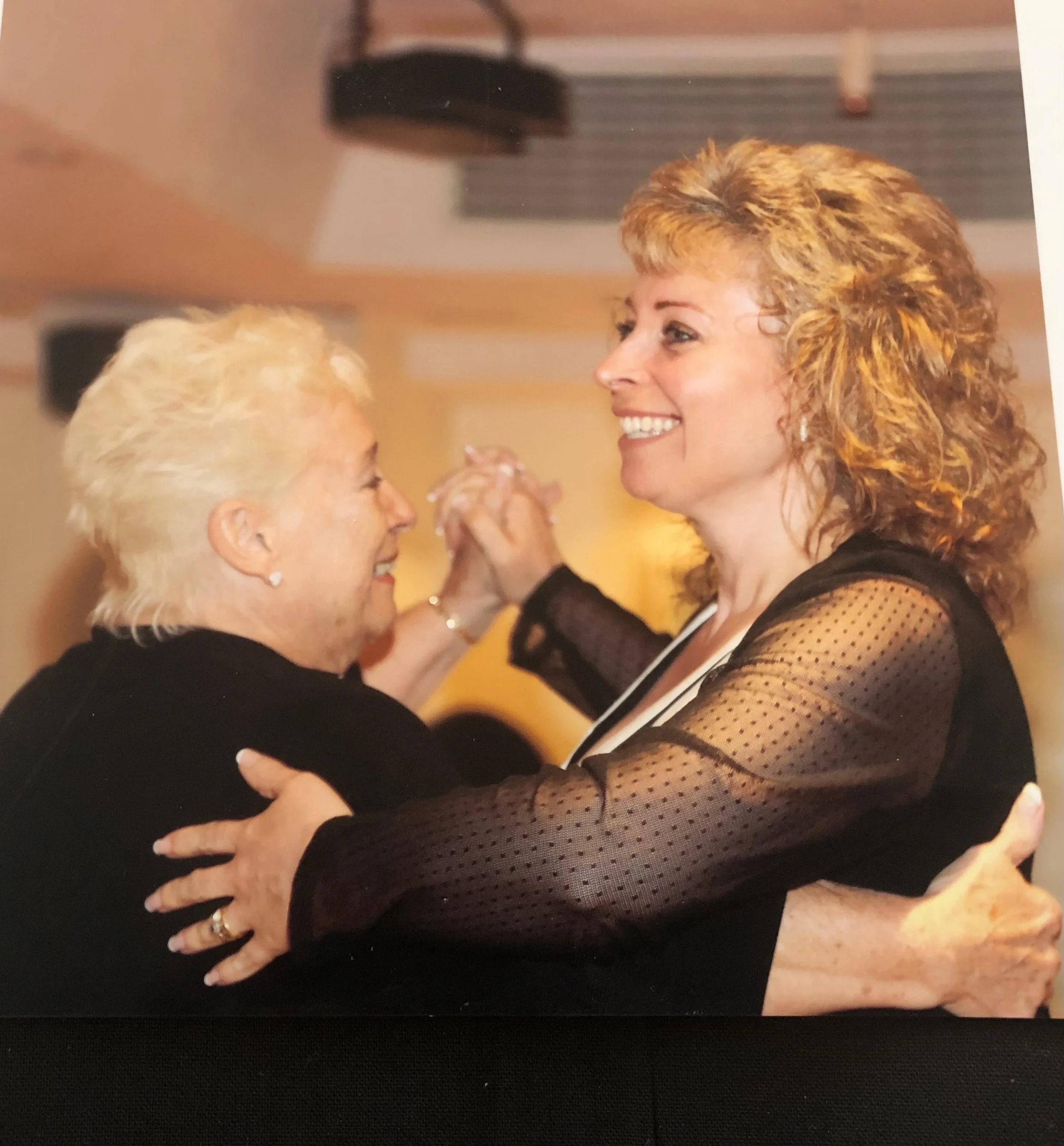 Two women dancing closely, smiling, and holding hands in a warm indoor setting.