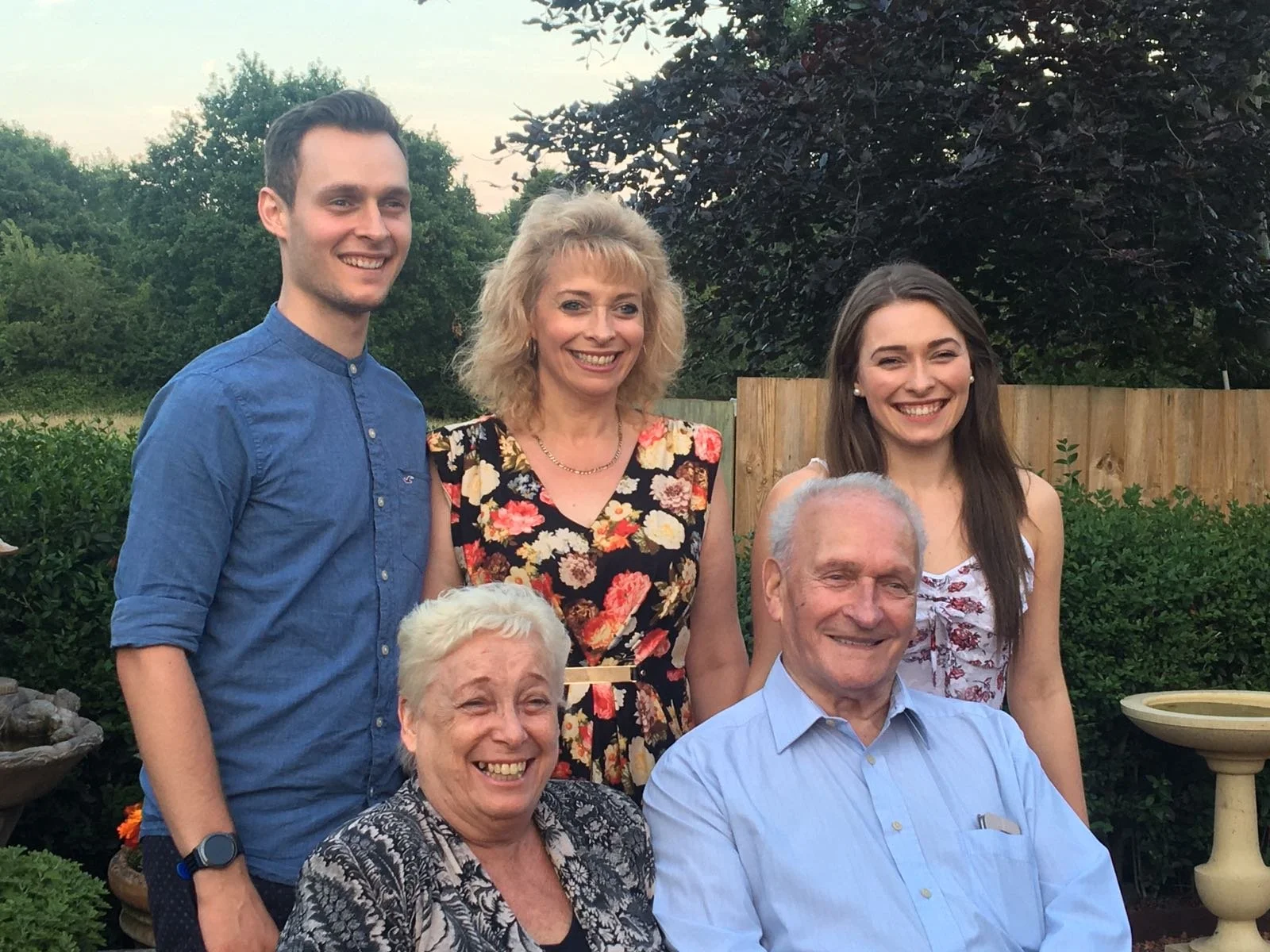 A family group photo outdoors with six people smiling. Two elderly people sitting in front, a middle-aged woman standing behind them, and three young adults standing behind her. The background shows trees, shrubs, and a wooden fence.