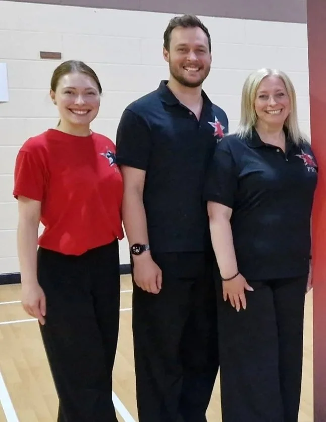 Three smiling people, two women and one man, standing indoors against a light-colored brick wall, wearing casual attire with logos, in a friendly group pose.