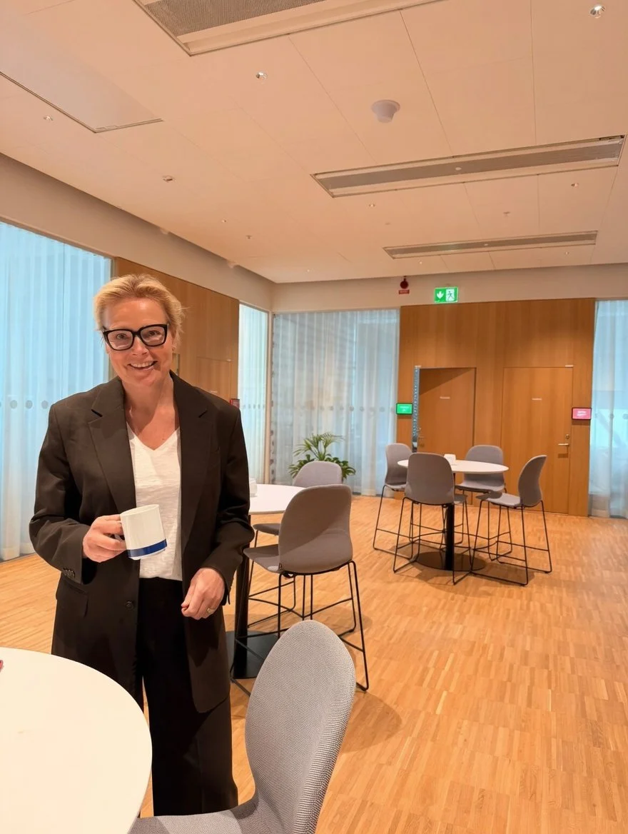 A woman with glasses and blonde hair, wearing a black blazer and white shirt, stands in a modern, well-lit room holding a white mug. The room has round tables and chairs, large windows with sheer curtains, wood-paneled walls, and a green exit sign.