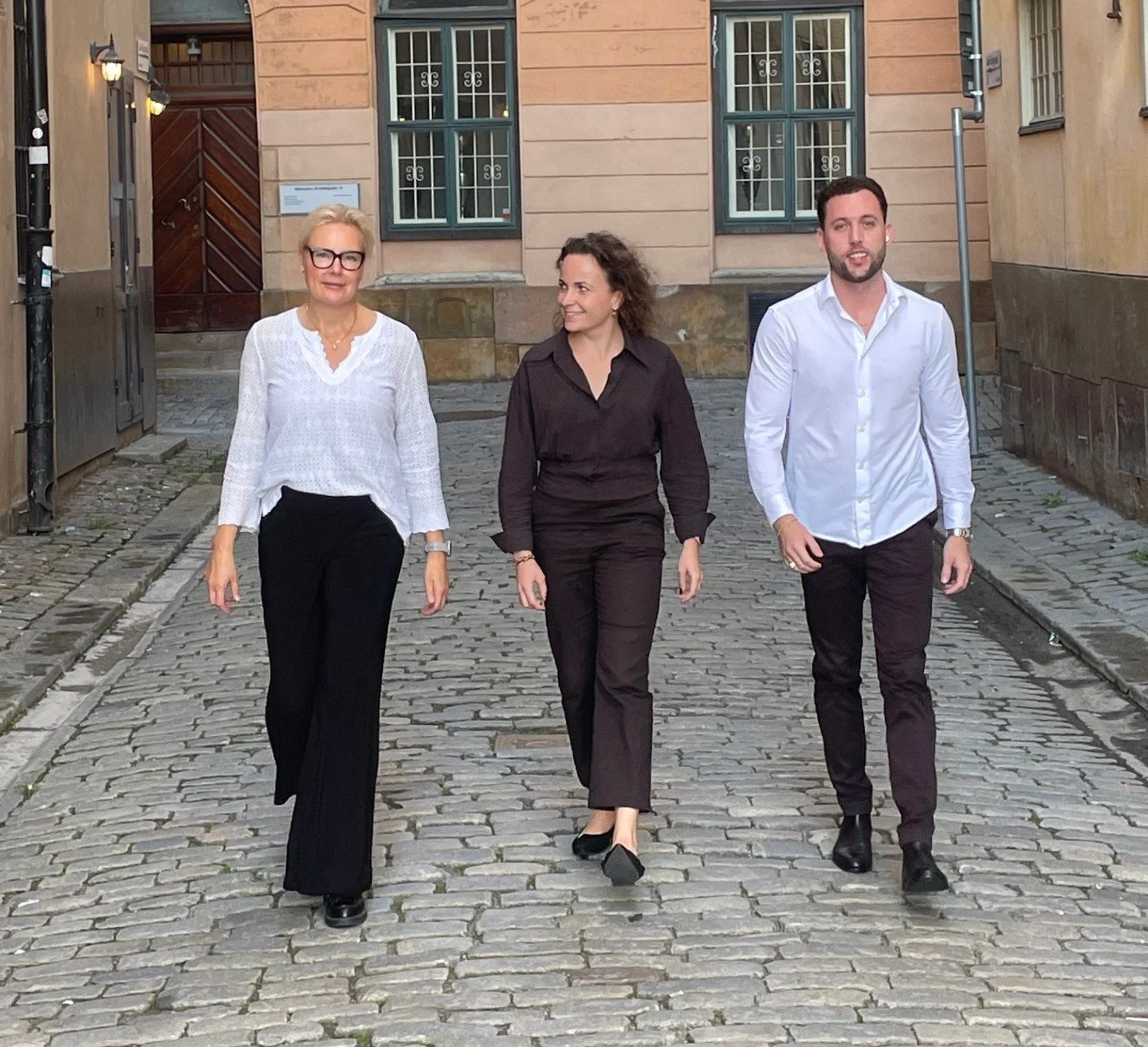 Three professionally dressed people walking on a cobblestone street in an urban area.