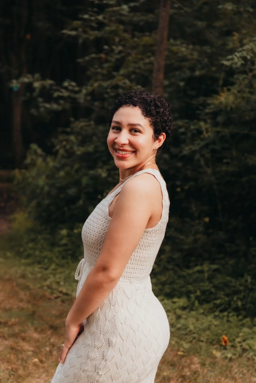 A smiling woman with short curly hair wearing a sleeveless cream dress outdoors with trees in the background.