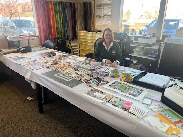 A woman sitting at a table covered with craft supplies, including colorful paper, tape, and printed materials, in a room with large windows and a view of parked cars outside.