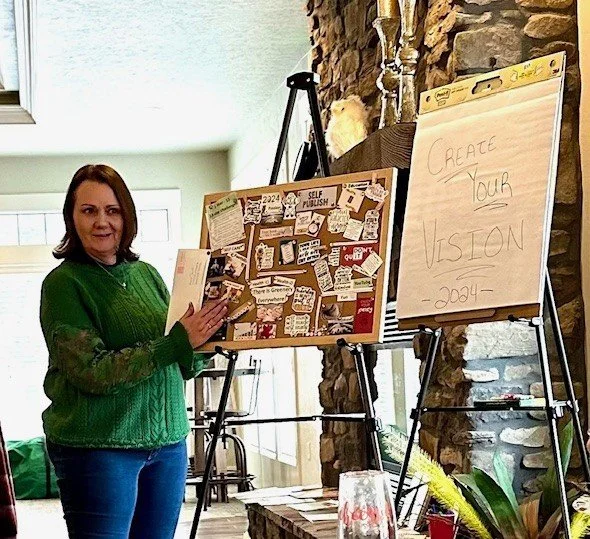 A woman standing next to a corkboard filled with various notes and images, holding a book, with a flip chart on an easel next to her that says 'Create Your Vision - 2024'.