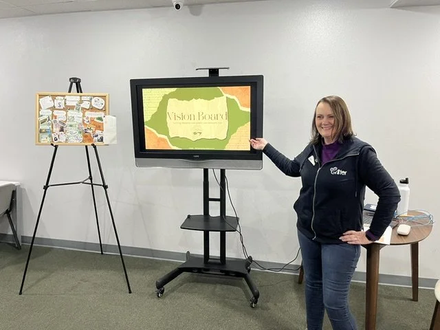 A woman standing in a conference room next to a large screen displaying a presentation titled 'Vision Board'. She is smiling and pointing at the screen, with a corkboard on a tripod stand to her left.