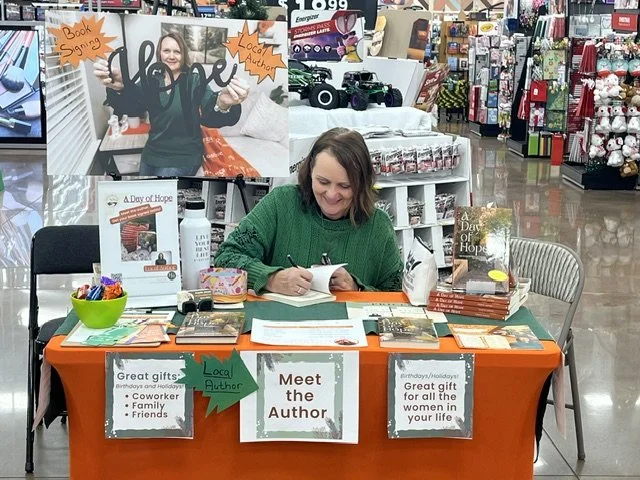 A woman sitting at a table in a store signing copies of her book, 'A Day of Hope.' The table has promotional signs, books, and a green bowl filled with candy, with shelving and holiday items in the background. Inset shows her standing with her arms outstretched, and signs indicating she's a local author.