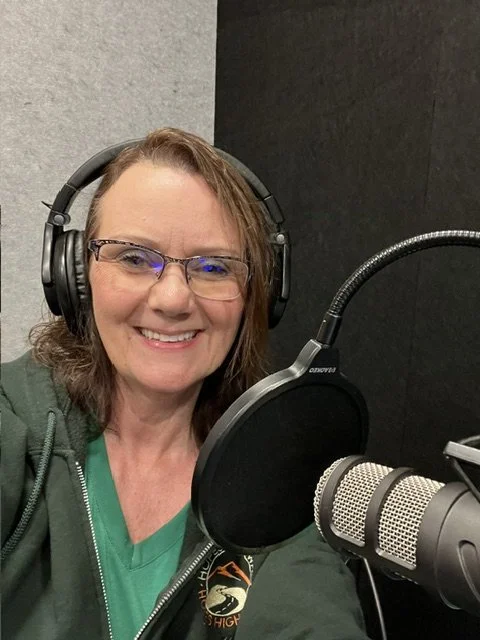 A woman with glasses and brown hair smiling while wearing headphones and sitting in front of a microphone in a recording studio.