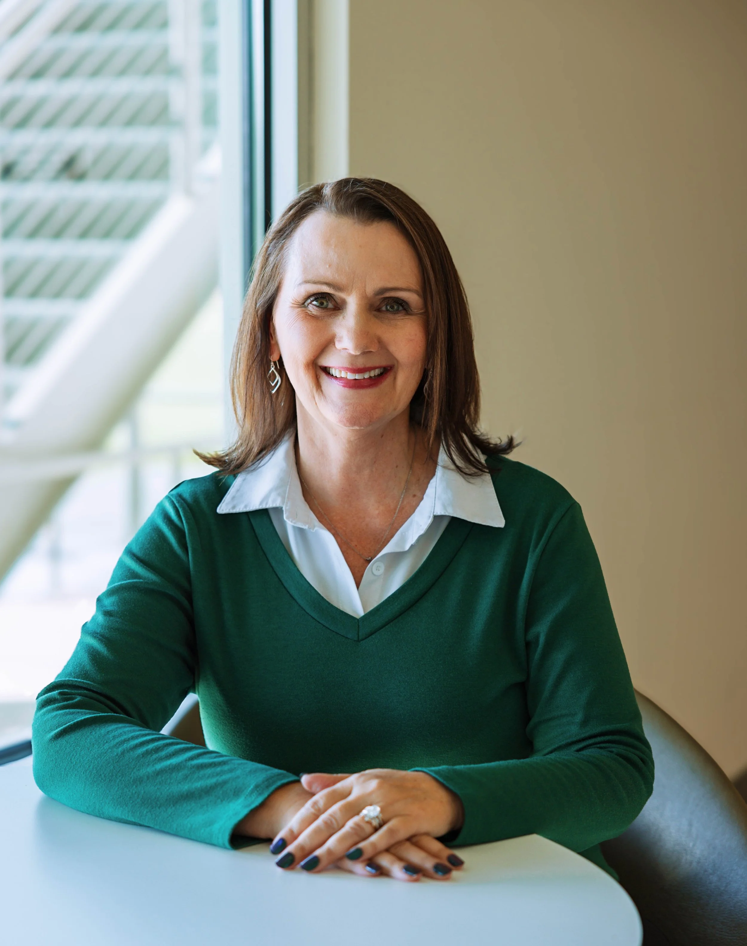 A woman with brown hair, green eyes, and fair skin smiling at the camera, sitting at a white table near a window in a well-lit room, wearing a green sweater over a white collared shirt, with her hands resting on the table.