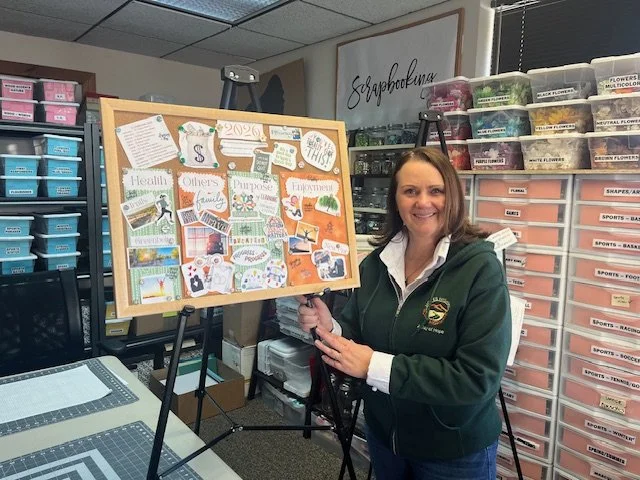 Woman standing next to a large bulletin board filled with colorful papers, in a craft or office supply room with storage drawers and containers around.