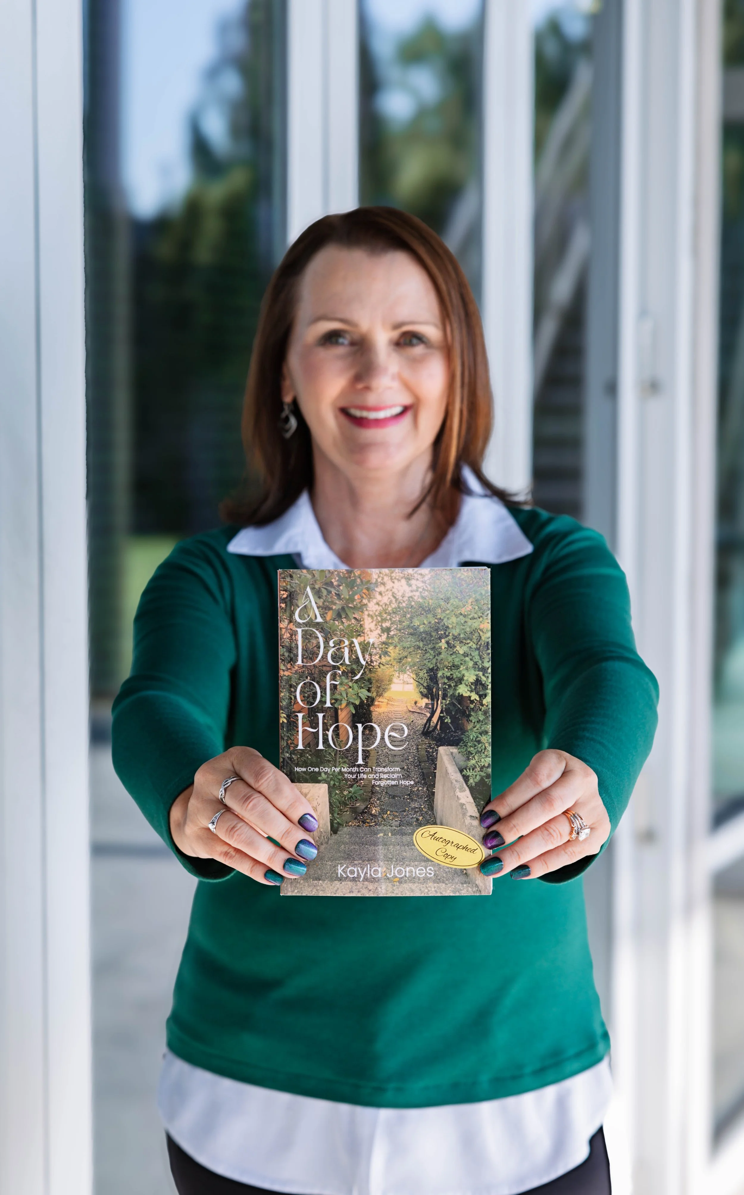 A woman with shoulder-length brown hair and a green sweater stands indoors, holding a book titled 'A Day of Hope'. The book cover features a peaceful path lined with trees. She is smiling and looking at the camera with painted fingernails.